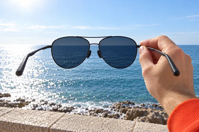 One hand holding the AceShades aviator sunglasses up against a bright sea, the water deep blue through the dark lenses