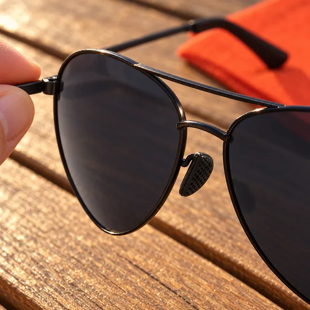 Close on the black double bridge, a nose pad and a dark lens of the AceShades, a hand holding the frame by its arm over a wooden table