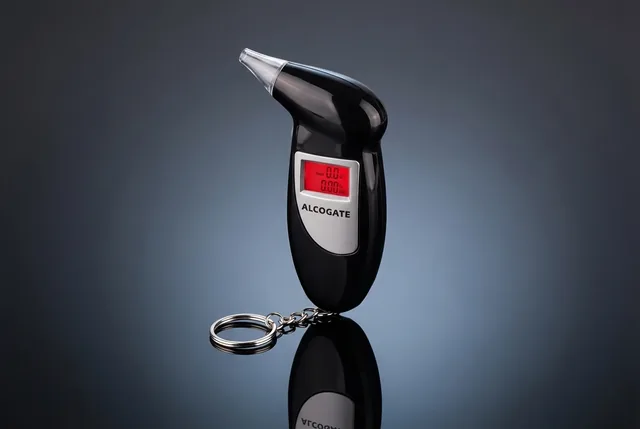 The black AlcoGate breathalyzer standing upright on a dark blue gray background, its red display lit and its key ring chain at the bottom