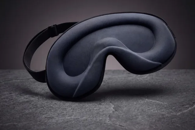 The BlackoutBliss sleep mask standing on its edge on dark slate, its gray molded inside facing out and its black elastic strap with the slide buckle behind it