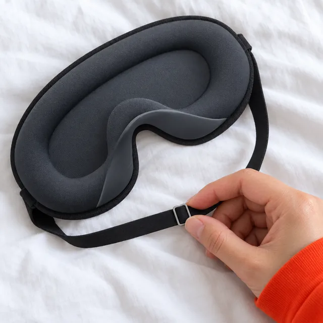 A hand moving the slide buckle on the black elastic strap of the charcoal gray BlackoutBliss mask, on white bed linen