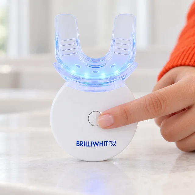 A fingertip pressing the round button in the middle of the white BrilliWhite unit, the clear mouthpiece above it lit blue
