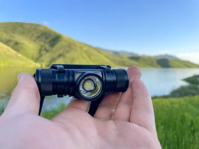 An open hand holding the Brillumex lamp in its cradle, the lens to the camera, a lake and green hills behind