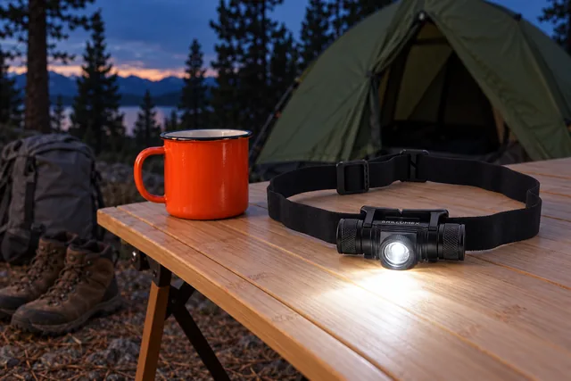 The Brillumex head torch on its headband lying on a camp table in front of a tent at dusk, its beam lit across the table beside a mug