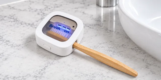 Das BrushSan-Etui, geschlossen über einer Bambuszahnbürste, auf einer Marmorablage neben dem Waschbecken, innen blau beleuchtet
