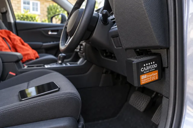 Der CarFixio steckt unter dem Armaturenbrett eines geparkten Autos, auf dem Fahrersitz liegt ein Handy
