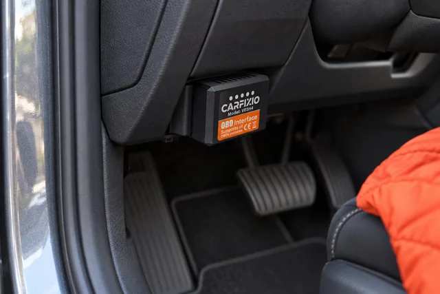 Der schwarze CarFixio steckt in der OBD2-Buchse unter dem Armaturenbrett eines Autos, das orangefarbene Etikett nach außen, darunter die Pedale