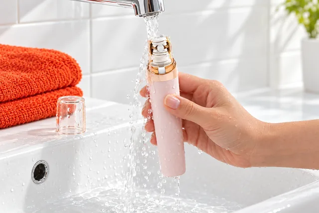 A hand holding the EpiGlow under a running tap at a white basin, its clear cap off and standing on the basin's edge