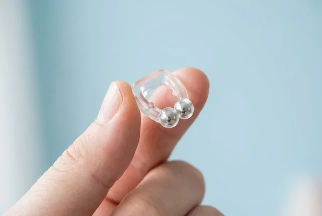 Two fingers holding the clear HushDream nose clip up against a pale blue background, its two silver spheres at the tips