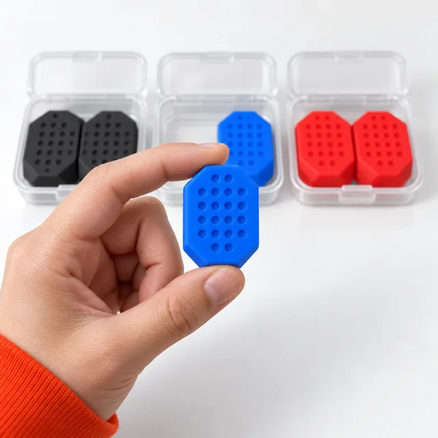 Fingers holding one blue JawCore trainer with its dimpled face to the camera, the black, blue and red pairs in their clear cases behind