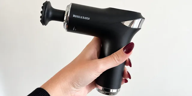 A hand holding the black MuscleAider by its grip against a white wall, the studded head on its chrome collar