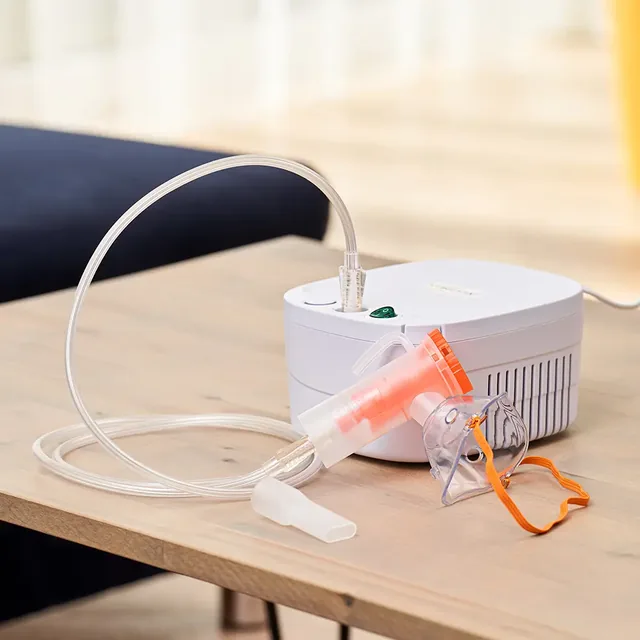 The white NebulaFix compressor on a light wooden table in front of a dark sofa, the clear tube running from it to the medicine cup, a clear mask with an orange strap fitted on the cup, the mouthpiece lying loose in front