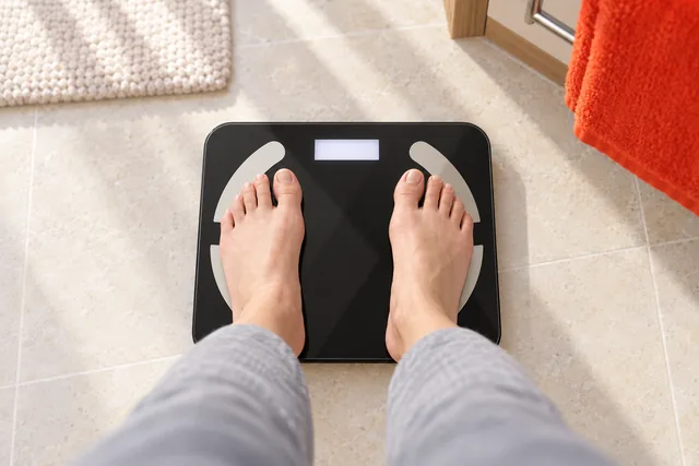 Two bare feet standing on the black ScanoPro scale on a pale bathroom floor, seen from above, its display lit