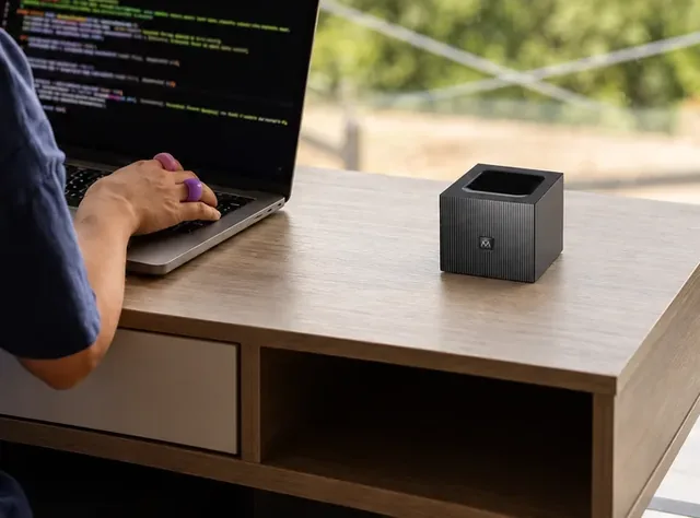 Der schwarze WifiQube auf einem Holzschreibtisch am Fenster, daneben tippt eine Hand auf einem Laptop